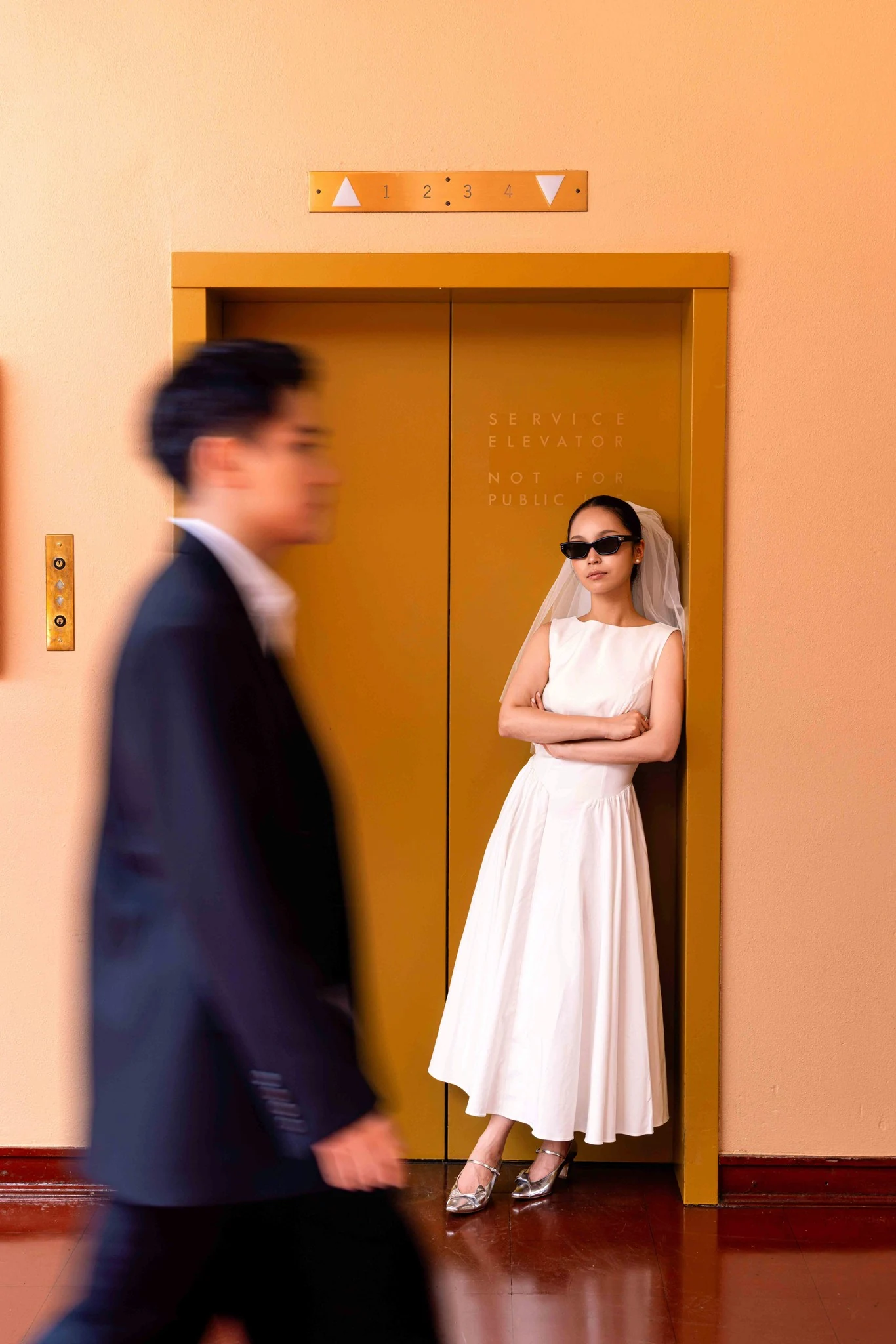 Bride in front of yellow elevator doors as the groom passes in the foreground